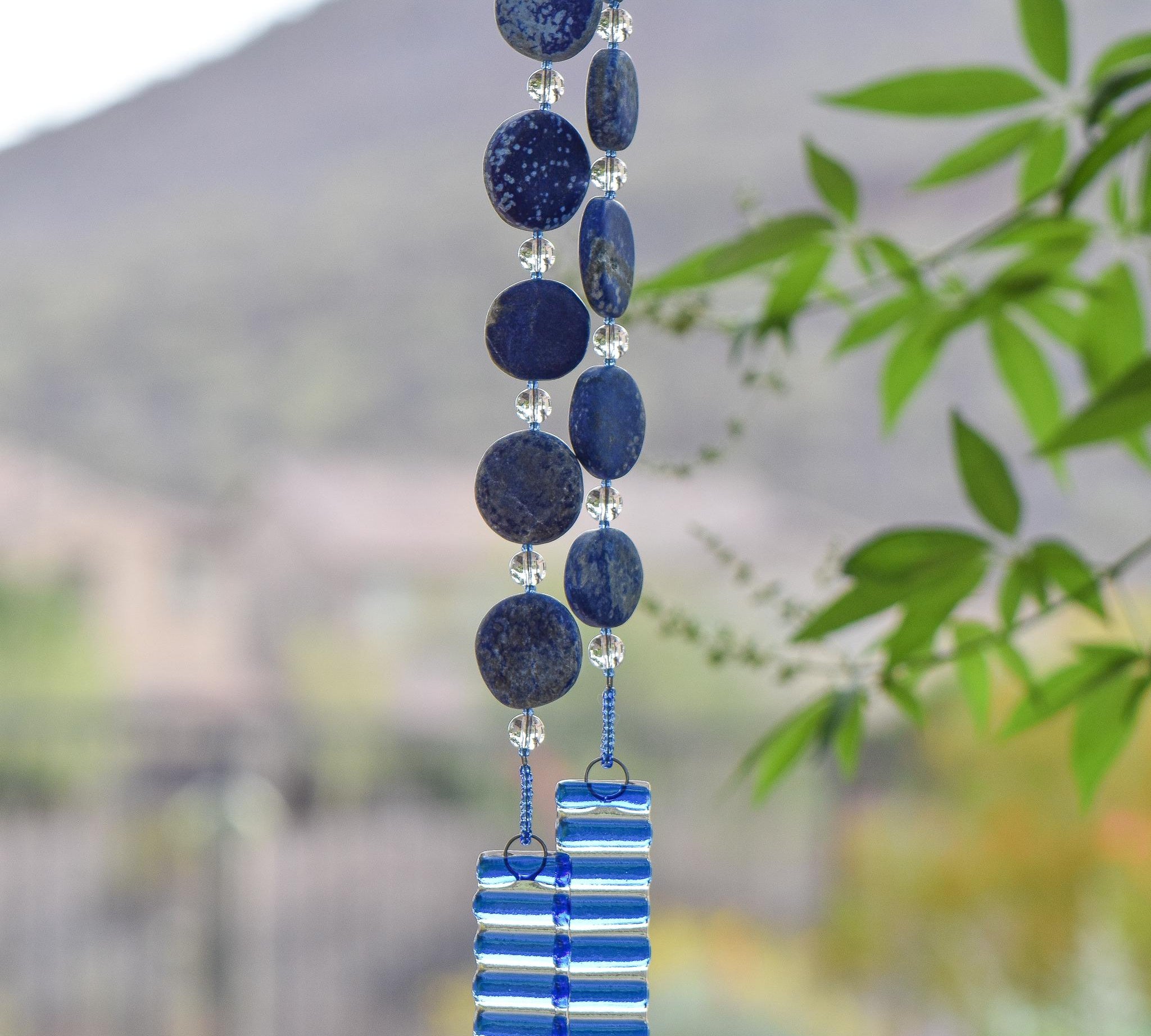 Hanging wind chime made with large thick lapis lazuli stone discs strung with glass beads, hanging vertically, anchored by two pieces of kiln formed clear glass with blue stripes.