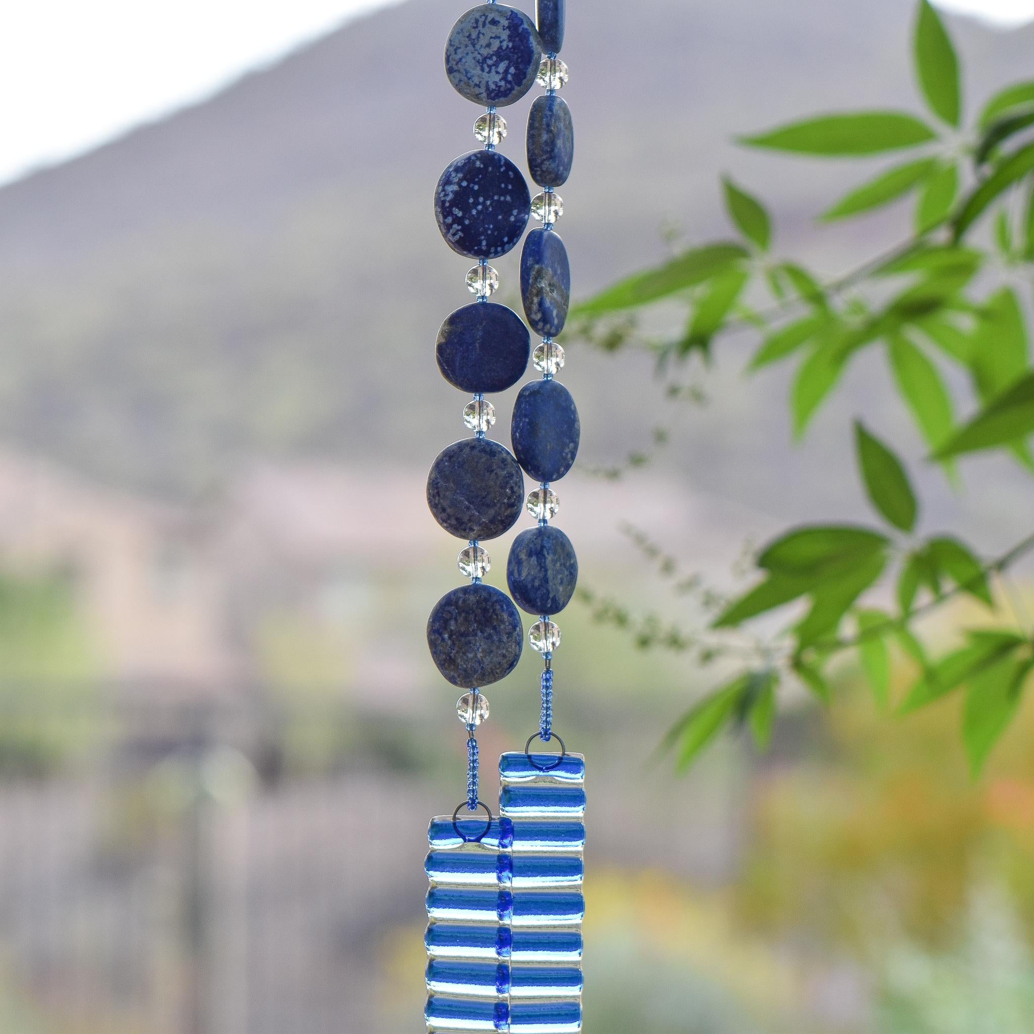 Hanging wind chime made with large thick lapis lazuli stone discs strung with glass beads, hanging vertically, anchored by two pieces of kiln formed clear glass with blue stripes.
