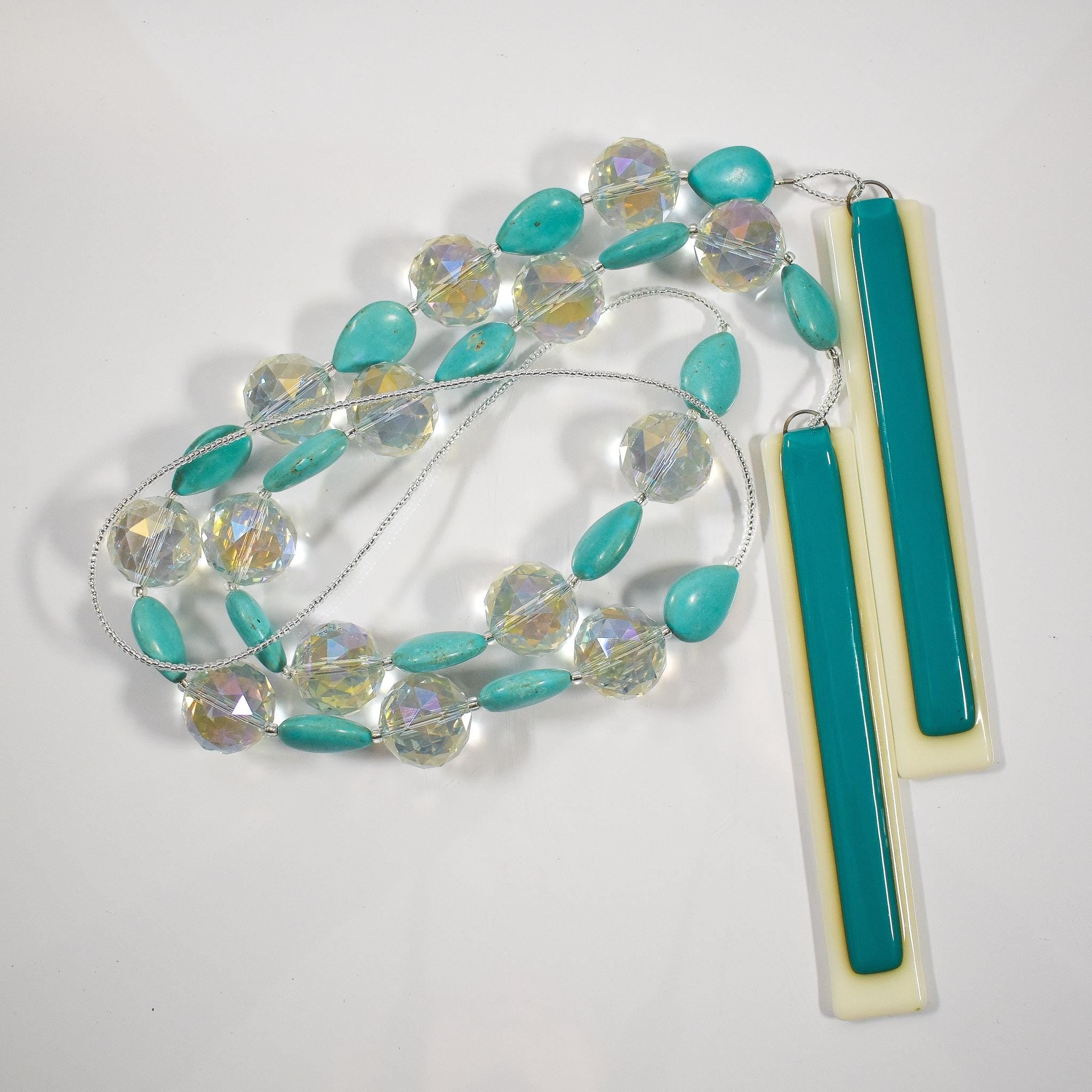 Turquoise Magnesite stone beads are teardrop-shaped and strung with large round clear glass crystal beads with lots of facets, anchored by two pieces of fused glass in vanilla cream and peacock green. Coiled against white background.