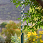 Turquoise Magnesite stone beads are teardrop-shaped and strung with large round clear glass crystal beads with lots of facets. All are hanging vertically against a mountain background, anchored by two pieces of kiln formed glass that is vanilla cream colored with a strip of peacock green.