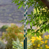 Turquoise Magnesite stone beads are teardrop-shaped and strung with large round clear glass crystal beads with lots of facets. All are hanging vertically against a mountain background, anchored by two pieces of kiln formed glass that is vanilla cream colored with a strip of peacock green.