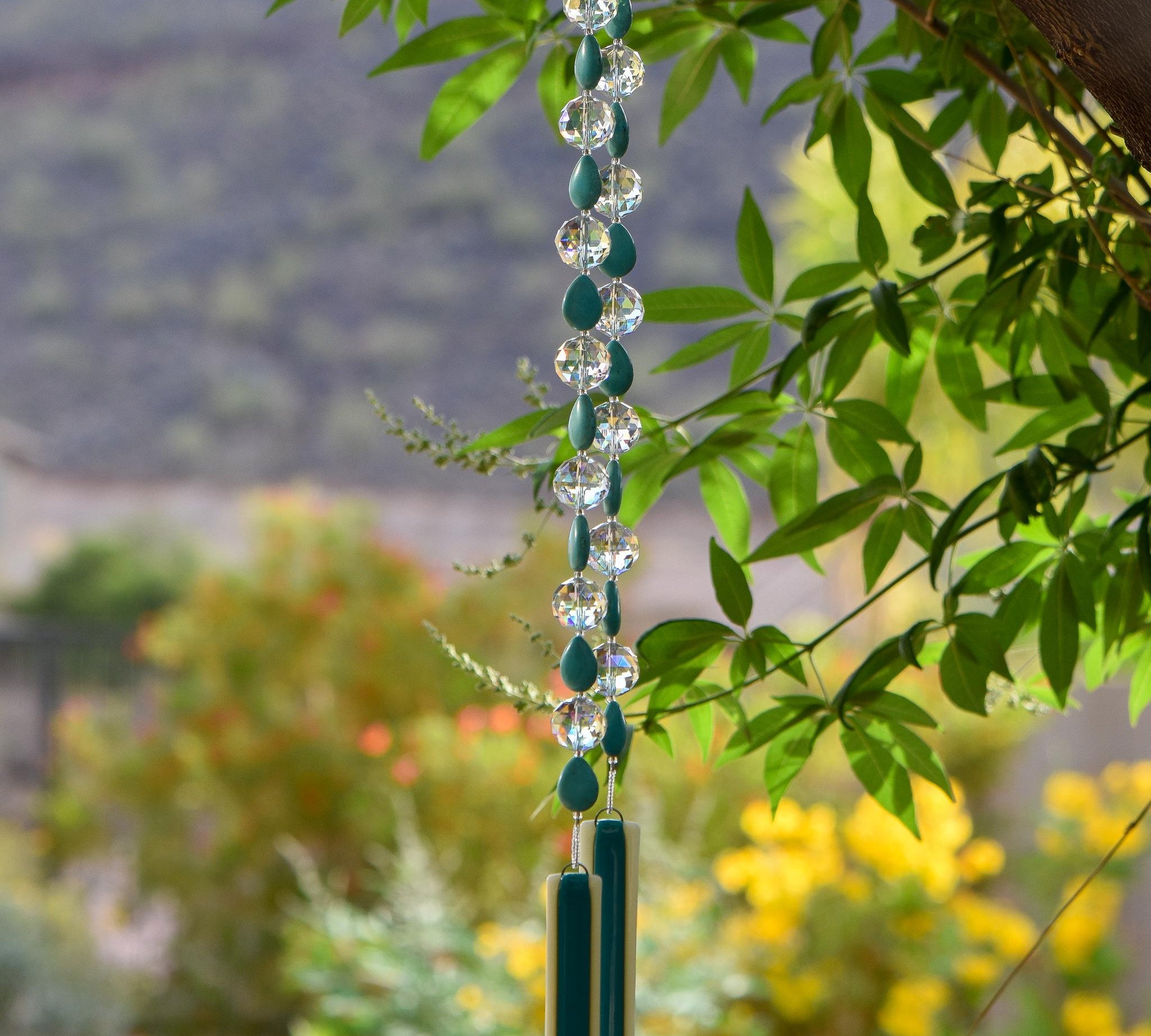 Turquoise Magnesite stone beads are teardrop-shaped and strung with large round clear glass crystal beads with lots of facets. All are hanging vertically against a mountain background, anchored by two pieces of kiln formed glass that is vanilla cream colored with a strip of peacock green.