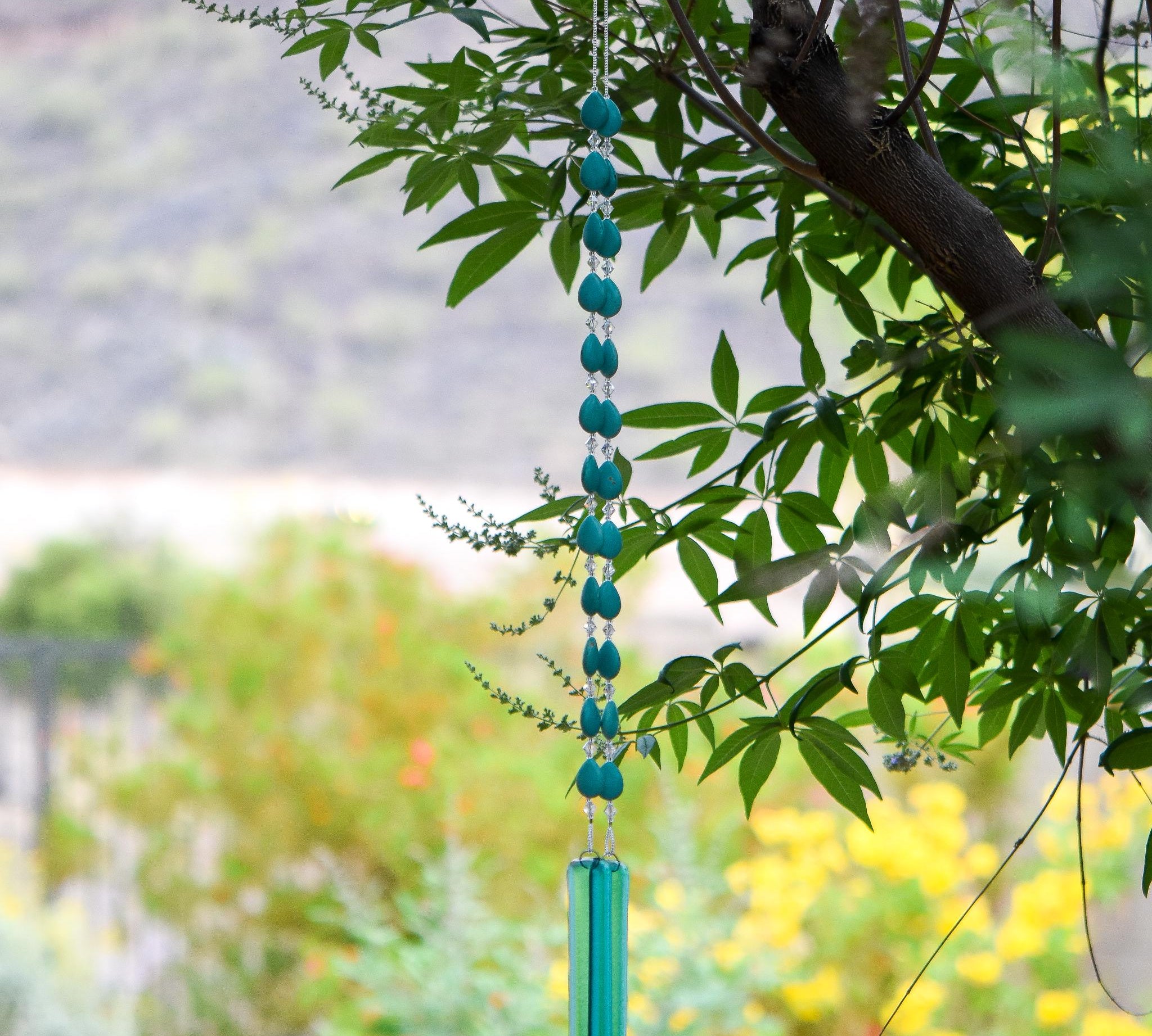 Long wind chime hanging from tree made with teardrop shaped magnesite stone beads, flanked by small glass crystals. Anchored by two pieces of transparent blue green glass.