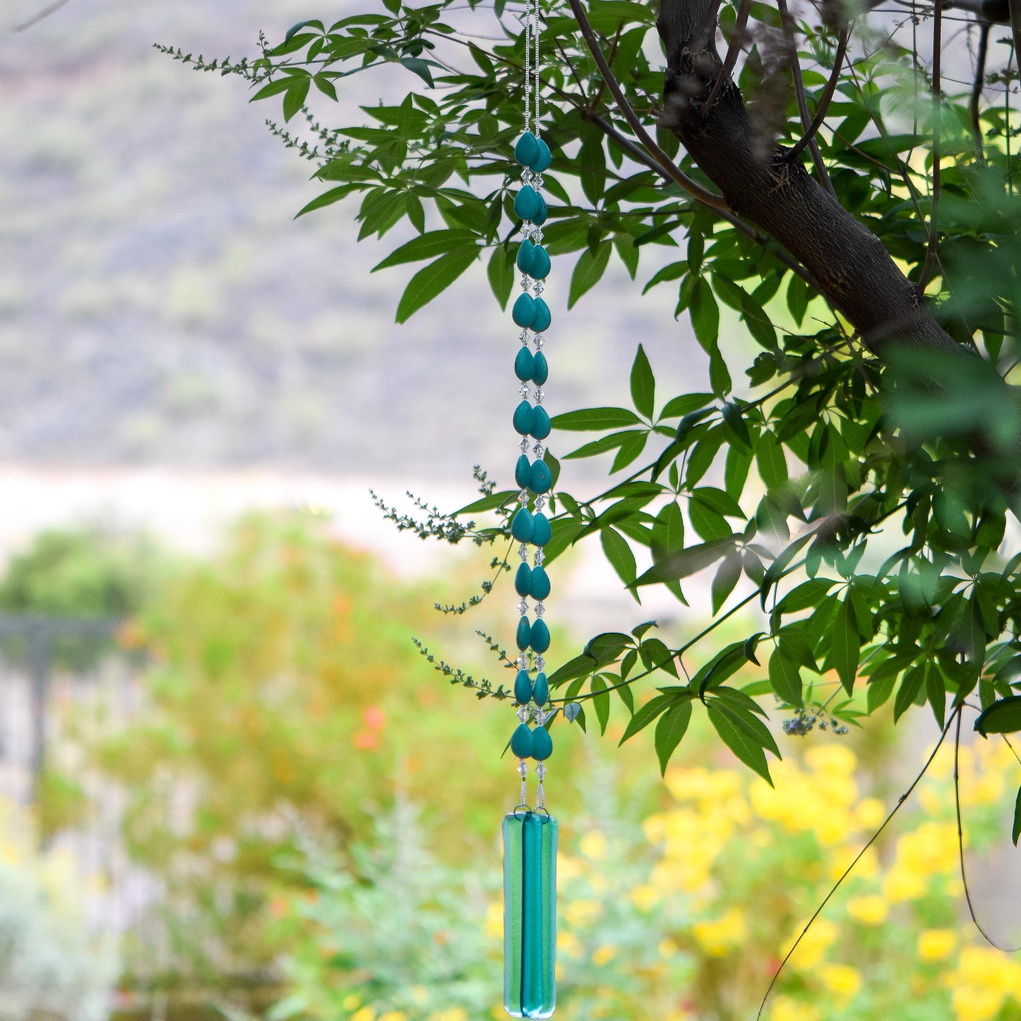 Long wind chime hanging from tree made with teardrop shaped magnesite stone beads, flanked by small glass crystals. Anchored by two pieces of transparent blue green glass.