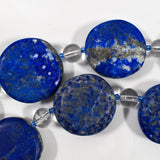 Large flat round lapis lazuli beads strung with smaller clear and blue glass beads, on white background.