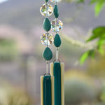 Turquoise Magnesite stone beads are teardrop-shaped and strung with large round clear glass crystal beads with lots of facets. All are hanging vertically against a mountain background, anchored by two pieces of kiln formed glass that is vanilla cream colored with a strip of peacock green.