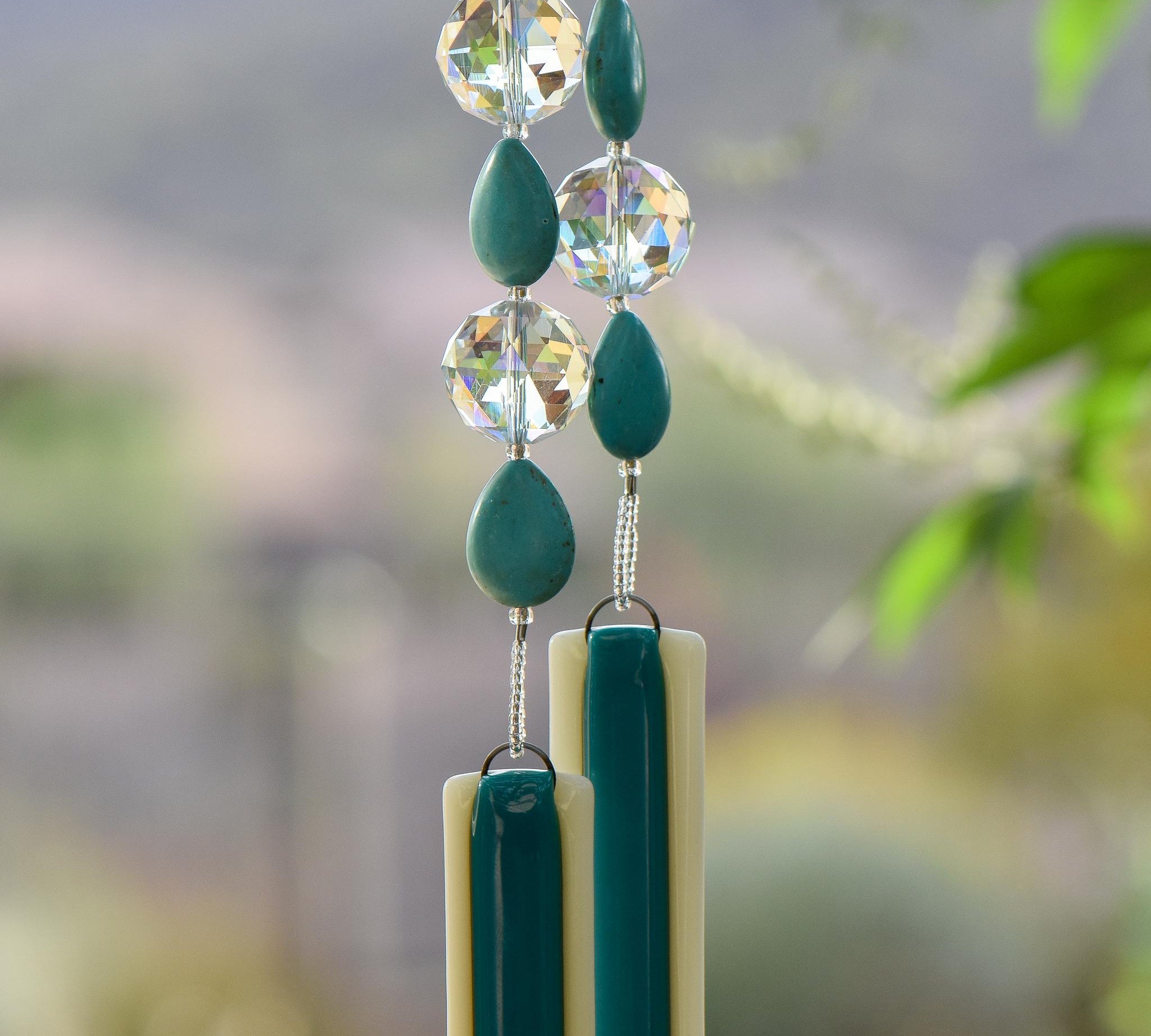 Turquoise Magnesite stone beads are teardrop-shaped and strung with large round clear glass crystal beads with lots of facets. All are hanging vertically against a mountain background, anchored by two pieces of kiln formed glass that is vanilla cream colored with a strip of peacock green.