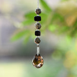 Small beaded sun catcher made with two black cube beads, three round tan beads with black spots and a large smoky quartz faceted tear drop pendant, hanging vertically against blurred background.