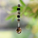 Small beaded sun catcher made with two black cube beads, three round tan beads with black spots and a large smoky quartz faceted tear drop pendant, hanging vertically against blurred background.
