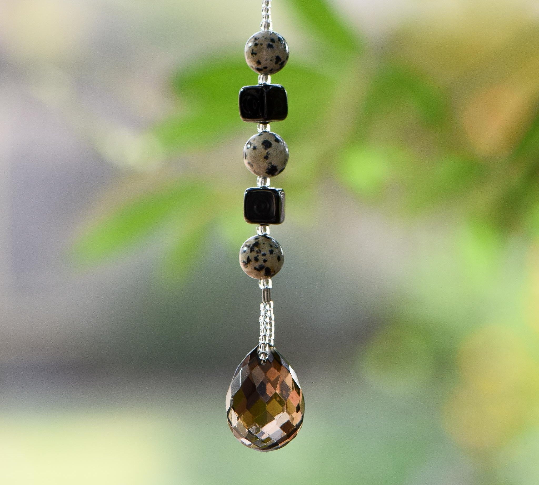 Small beaded sun catcher made with two black cube beads, three round tan beads with black spots and a large smoky quartz faceted tear drop pendant, hanging vertically against blurred background.