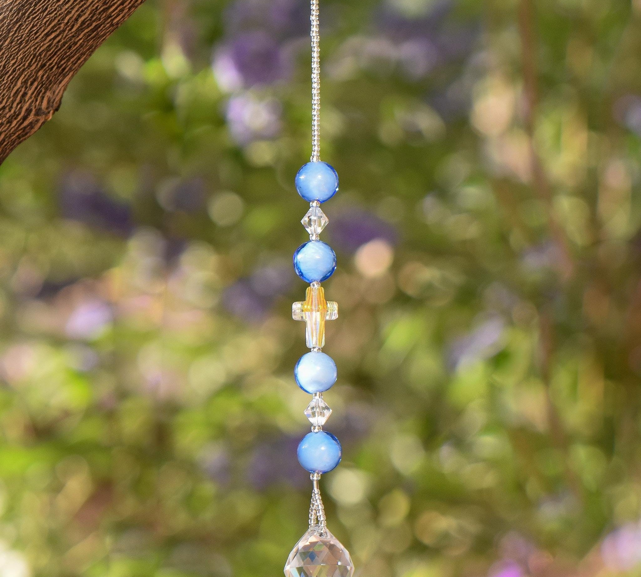 Beaded sun catcher hanging vertically against blurred green foliage background. Sun catcher has 4 light blue round glass beads, two bicone crystal beads. Center is a faceted glass cross bead with Crystal AB finish. Anchored by faceted glass round prism.