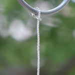 May include: A close-up shot of a wire covered in silver-lined seed beads  hanging from a silver ring. The ring is smooth and circular, and the chain is attached to it. The background is a blurred green, suggesting an outdoor setting.