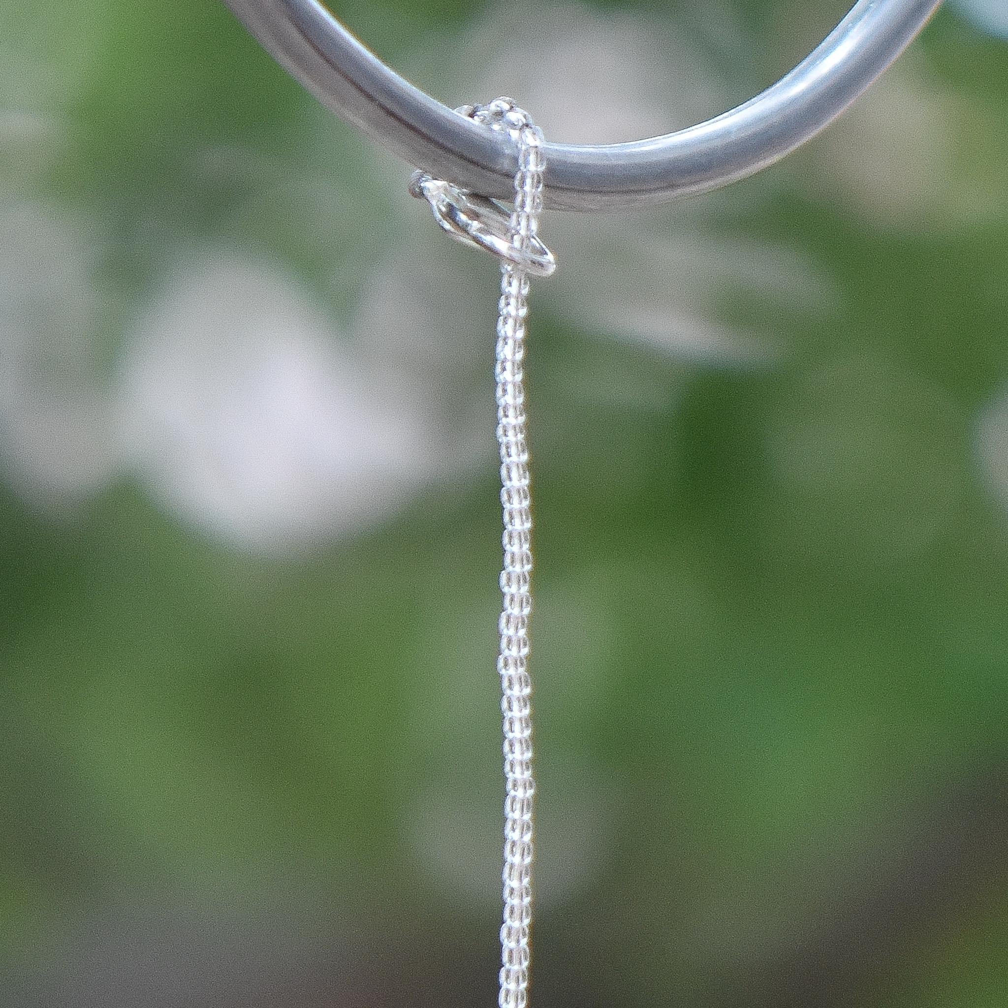 May include: A close-up shot of a wire covered in silver-lined seed beads  hanging from a silver ring. The ring is smooth and circular, and the chain is attached to it. The background is a blurred green, suggesting an outdoor setting.