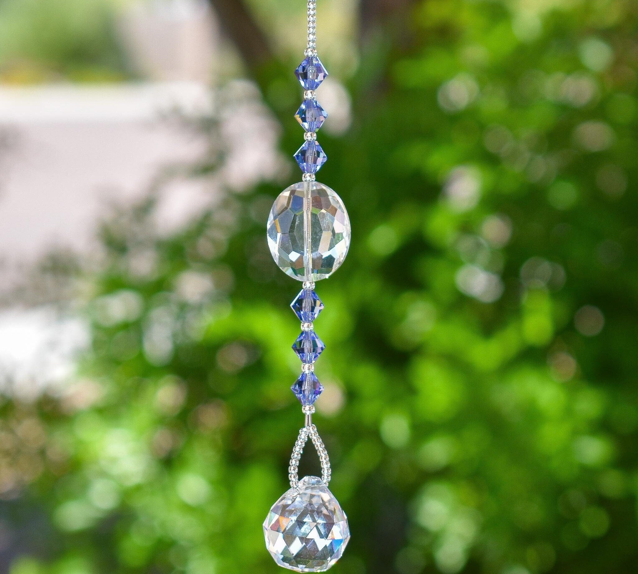 Large oval crystal bead flanked by six bicones in tanzanite color, anchored by round crystal prism, hanging vertically from tree.