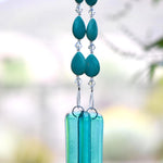 Teardrop-shaped turquoise magnesite stone beads strung with smaller clear glass crystal beads, hanging vertically, anchored by two pieces of transparent blue green glass against blurred background.