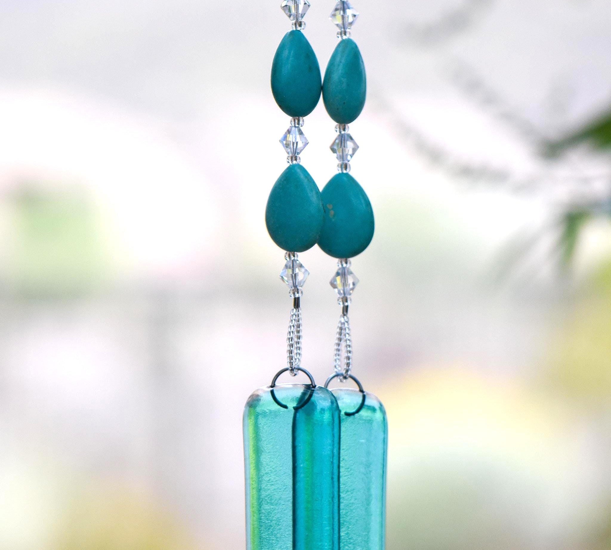 Teardrop-shaped turquoise magnesite stone beads strung with smaller clear glass crystal beads, hanging vertically, anchored by two pieces of transparent blue green glass against blurred background.