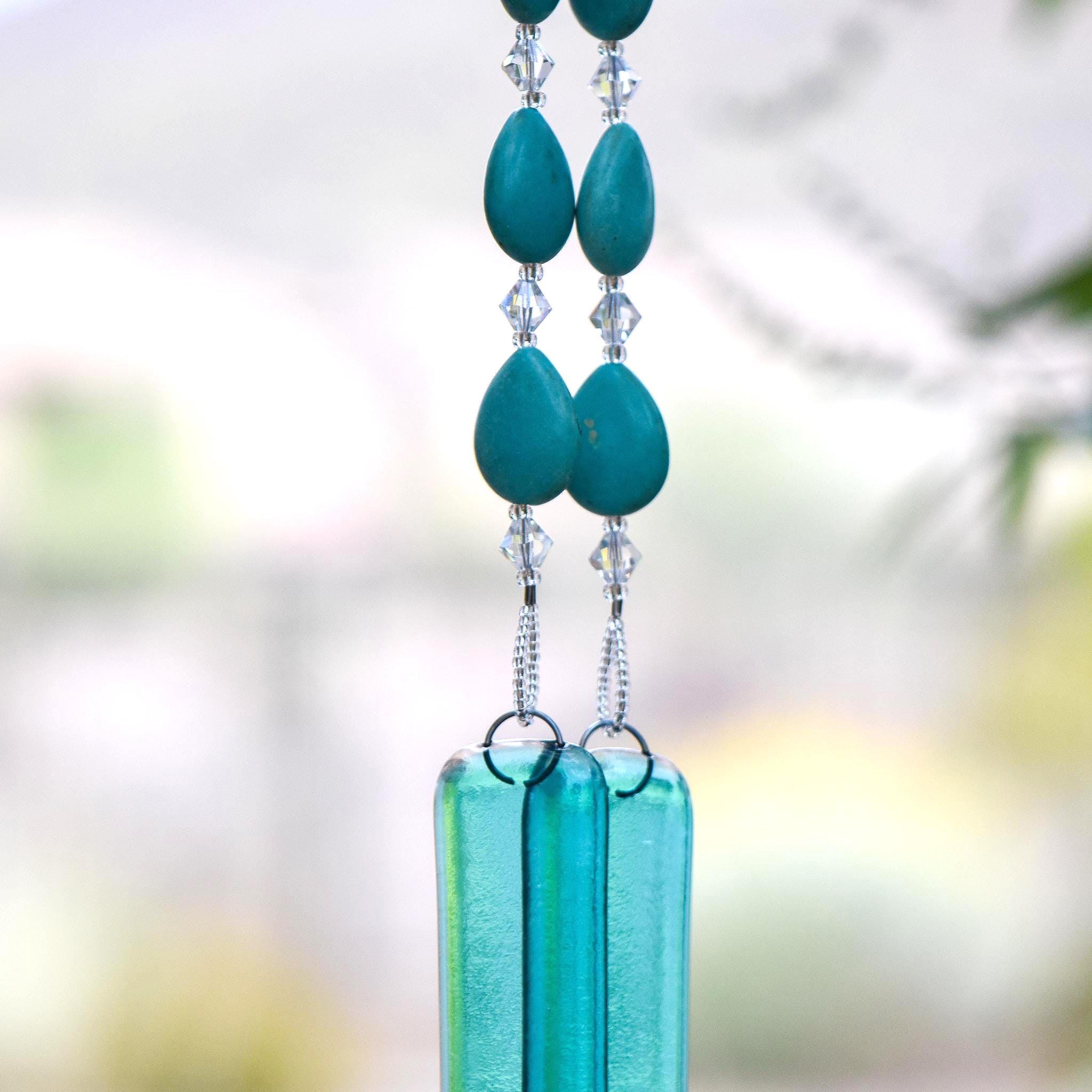 Teardrop-shaped turquoise magnesite stone beads strung with smaller clear glass crystal beads, hanging vertically, anchored by two pieces of transparent blue green glass against blurred background.