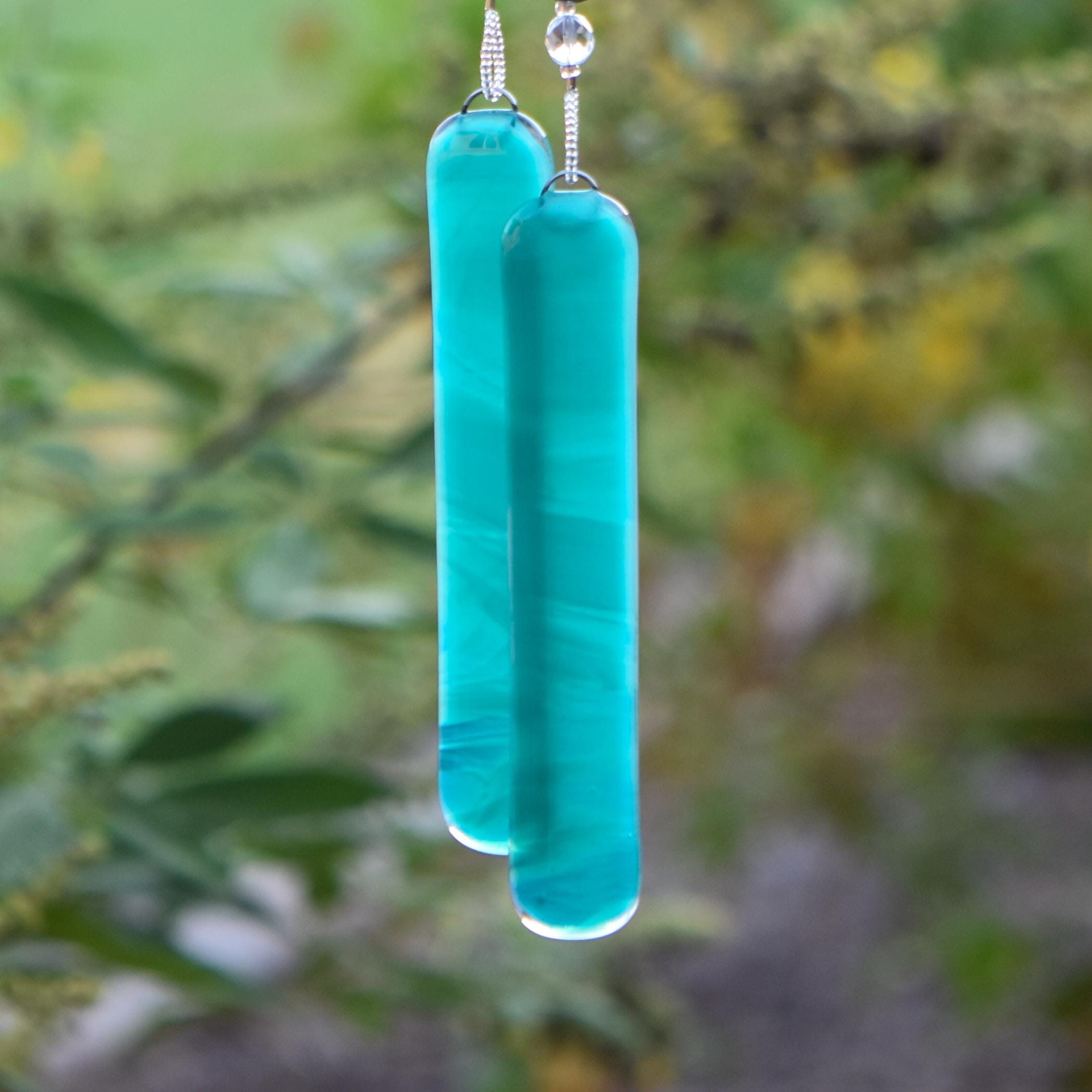 Two teal green rectangular pieces of glass attached to wire covered in silver-lined seed beads. The glass is streaked with white. The background is blurred foliage.