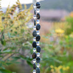 Two decorative beaded strands featuring alternating dark oval-shaped beads and clear round beads. The dark beads have a mottled appearance, and the clear beads are interspersed between them. The beads are hanging together, creating a vertical design, as part of a wind chime. The background is blurred, with green foliage and hints of other colors.