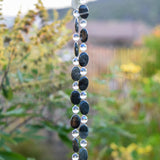 Two decorative beaded strands featuring alternating dark oval-shaped beads and clear round beads. The dark beads have a mottled appearance, and the clear beads are interspersed between them. The beads are hanging together, creating a vertical design, as part of a wind chime. The background is blurred, with green foliage and hints of other colors.