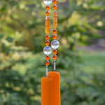 Acrylic and glass bright orange beads are strung on a wire with large round clear glass beads. The wire hangs vertically against a blurred foliage background. Wire is anchored by two rectangular pieces of opaque orange kiln formed glass.
