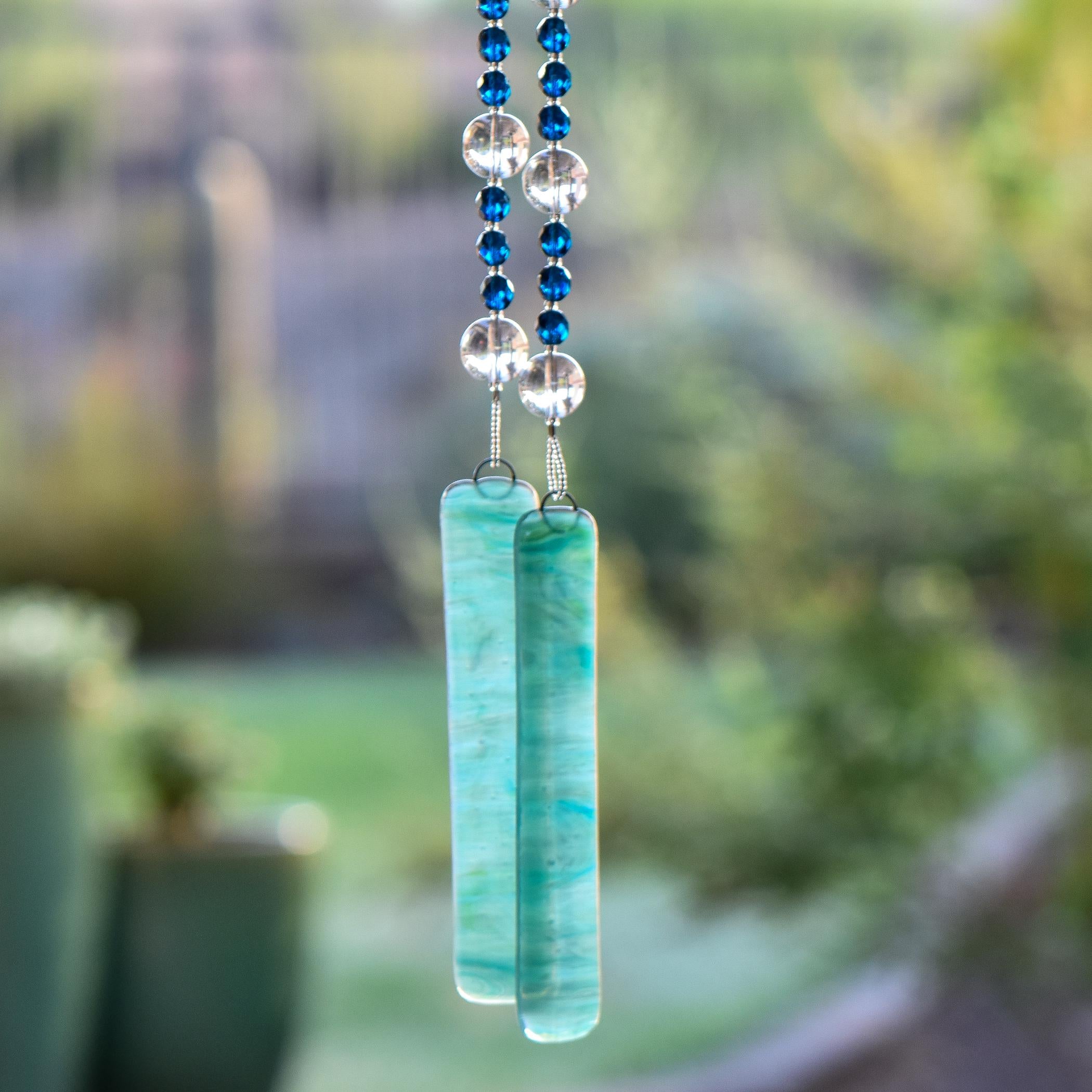 Dark Aqua Blue glass beads are strung vertically with larger clear glass beads, hanging against blurred landscape background. There are two pieces of lime green and aqua blue fused glass that anchor each end of the string. It is hung so that the glass can touch and create the sound.