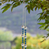 Dark Aqua Blue glass beads are strung vertically with larger clear glass beads, hanging against blurred landscape background. This photo shows the hanging point where the wire is draped over a hook that is hanging from a tree.