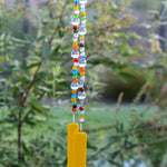 Multi-color glass beads from the Krobo tribe in Ghana, Africa strung with larger clear glass beads, hanging vertically in outdoor setting. Two strings of beads are anchored by two pieces of sunflower yellow fused glass.