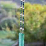 Dark Aqua Blue glass beads are strung vertically with larger clear glass beads, hanging against blurred landscape background. There are two pieces of lime green and aqua blue fused glass that anchor each end of the string. It is hung so that the glass can touch and create the sound.