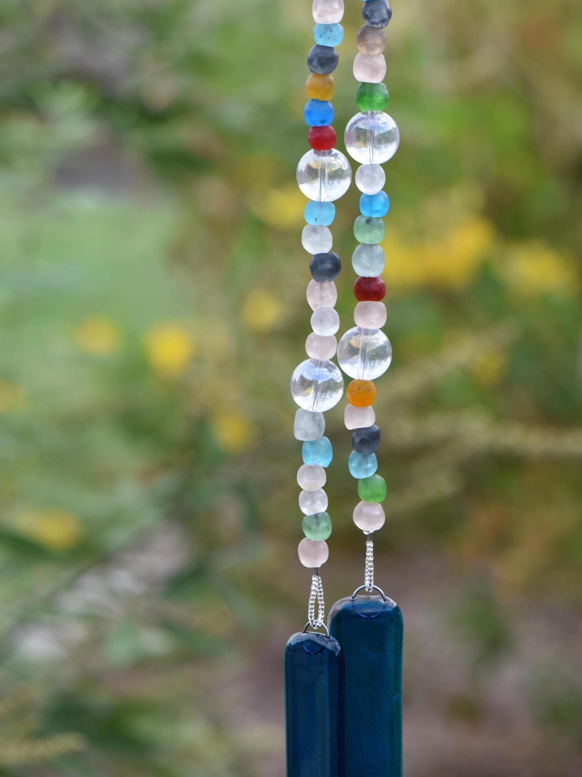 Brightly colored glass beads from Ghana, South Africa&#39;s Krobo tribe are strung with large solid clear glass beads, hanging in front of blurred foliage background. Two pieces of deep aqua blue fused glass are at each end of the long wire.