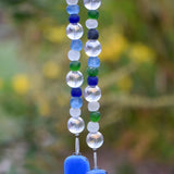 Handmade glass wind chime featuring alternating round and frosted beads in shades of blue, green, clear, and white. The beads are strung vertically and end in two smooth cobalt blue glass pieces, with a softly blurred garden background in green and yellow tones.