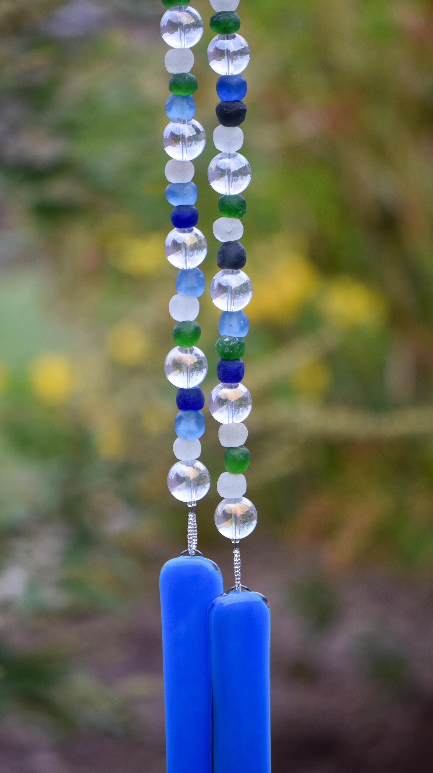 Handmade glass wind chime featuring alternating round and frosted beads in shades of blue, green, clear, and white. The beads are strung vertically and end in two smooth cobalt blue glass pieces, with a softly blurred garden background in green and yellow tones.
