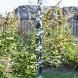 Two decorative beaded strands featuring alternating oval-shaped, speckled, dark blue stones and clear, round beads. Small, blue beads separate the larger stones and clear beads. The strands hang vertically against a blurred background of green foliage and a fence.