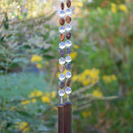 Bottom of Cinjo Handmade wind chime hanging in front of blurred foliage background. Two piece of transparent pale purple fused glass, rectangular-shaped, are anchors for two strings of beaded wire. Rhyolite stone beads are tear drop shaped and there is a larger, clear, reflective glass bead in between each stone bead. The stone beads are marked and in shades of brown, tan and gray.