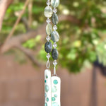 Large oval green quartz stone beads are strung with smaller clear glass beads, hanging vertically from a tree. Two fused glass pieces in vanilla cream with transparent teal embellishments provide the anchor for this unique handmade chime.