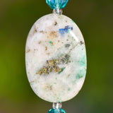 Close up view of large, oval stone azurite malachite stone bead against blurred green background. Stone is mostly white with some inclusions and some green veining.