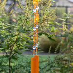 Acrylic and glass bright orange beads are strung on a wire with large round clear glass beads. The wire hangs vertically against a blurred foliage background. Wire is anchored by two rectangular pieces of opaque orange kiln formed glass.
