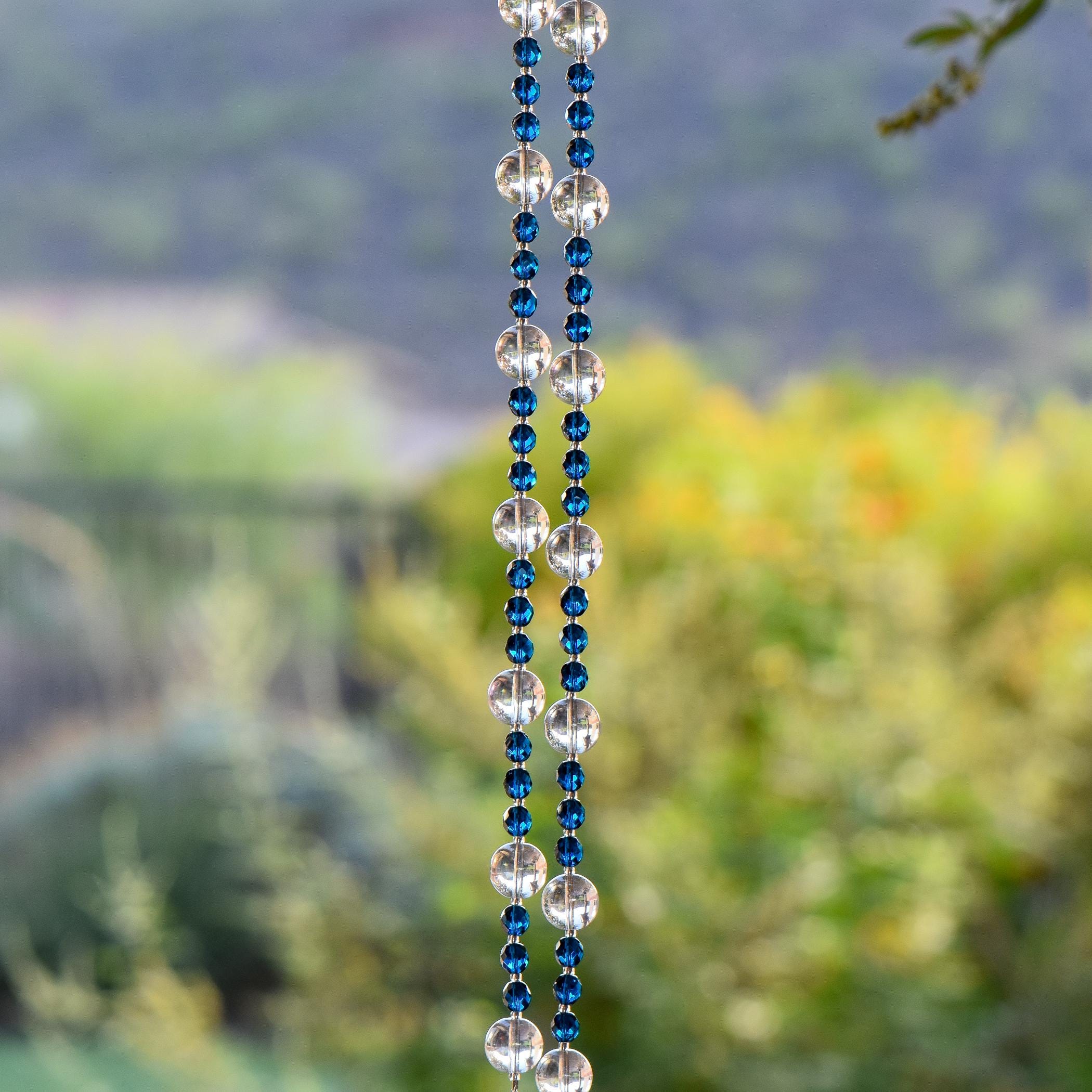 Dark Aqua Blue glass beads are strung vertically with larger clear glass beads, hanging against blurred landscape background. The piece is draped over a hook so that there are two legs that hang down.