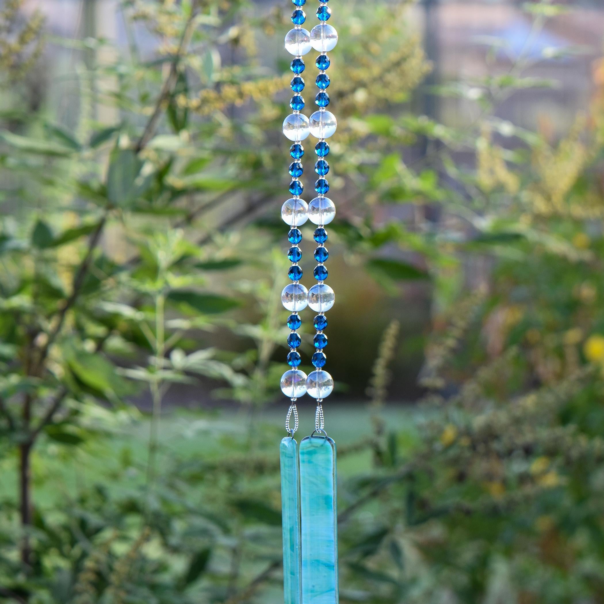 Dark Aqua Blue glass beads are strung vertically with larger clear glass beads, hanging against blurred landscape background. There are two pieces of lime green and aqua blue fused glass that anchor each end of the string. It is hung so that the glass can touch and create the sound.