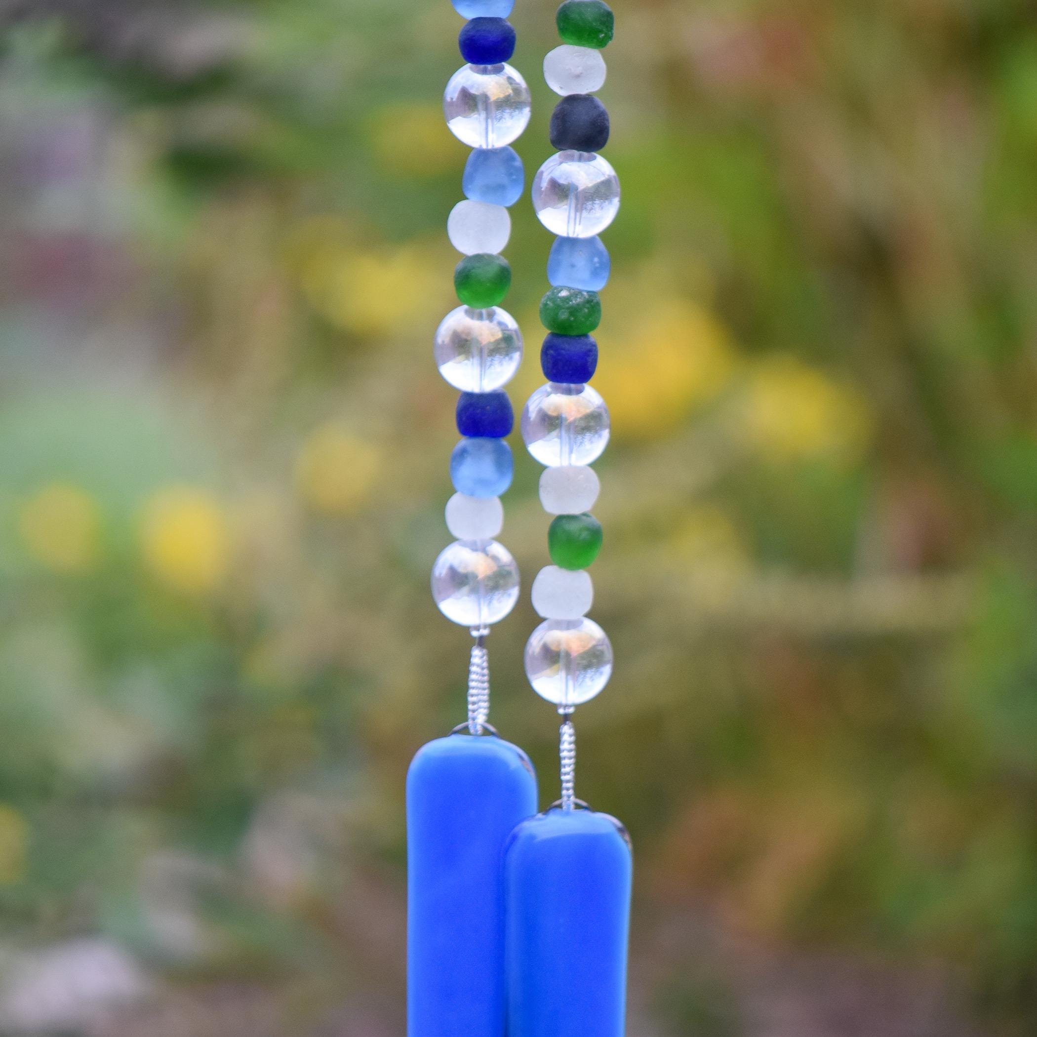 Close-up of a handmade glass wind chime featuring alternating round and frosted beads in shades of blue, green, clear, and white. The beads are strung vertically and end in two smooth cobalt blue glass pieces, with a softly blurred garden background in green and yellow tones.