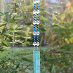 Dark Aqua Blue glass beads are strung vertically with larger clear glass beads, hanging against blurred landscape background. There are two pieces of lime green and aqua blue fused glass that anchor each end of the string. It is hung so that the glass can touch and create the sound.