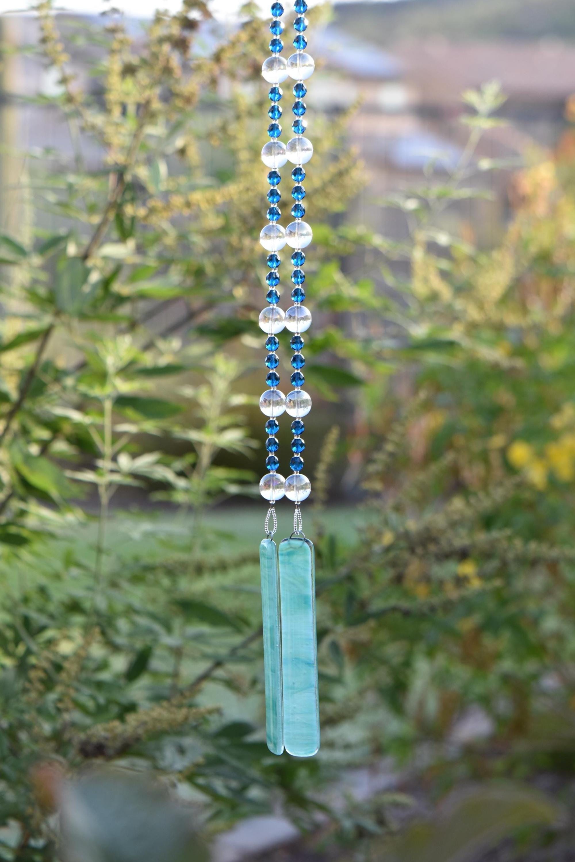 Dark Aqua Blue glass beads are strung vertically with larger clear glass beads, hanging against blurred landscape background. There are two pieces of lime green and aqua blue fused glass that anchor each end of the string. It is hung so that the glass can touch and create the sound.