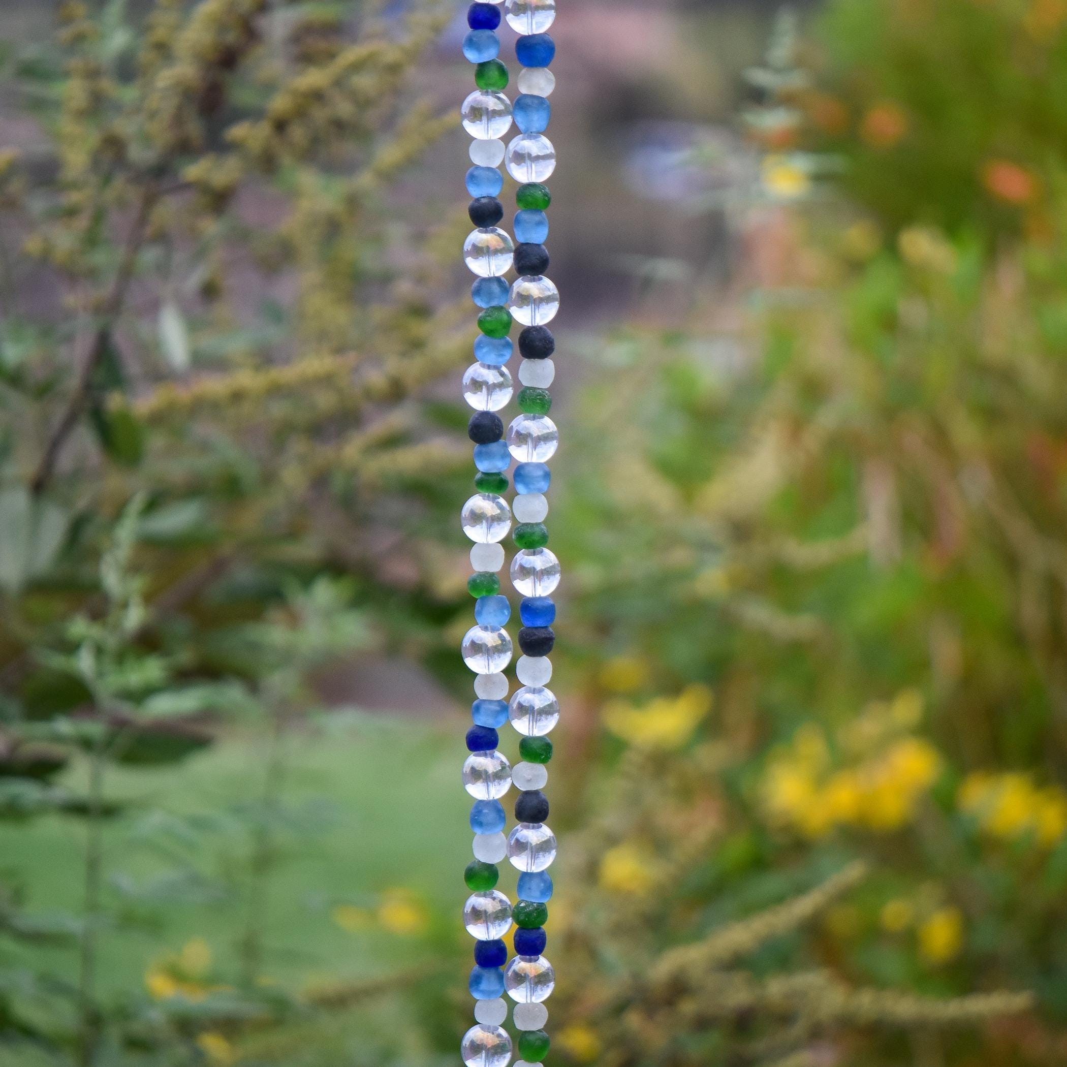A beaded wind chime with a variety of colorful beads. The beads are primarily clear, blue, and green, arranged in a vertical pattern. The beads are round and transparent, creating a decorative and eye-catching design. The wind chime is used for outdoor decoration.