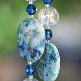 Close-up of a beaded wind chime featuring large, oval-shaped stones with a blue and green marbled pattern. The stones are interspersed with faceted, deep blue beads and clear, round beads. The background is a soft, blurred green.