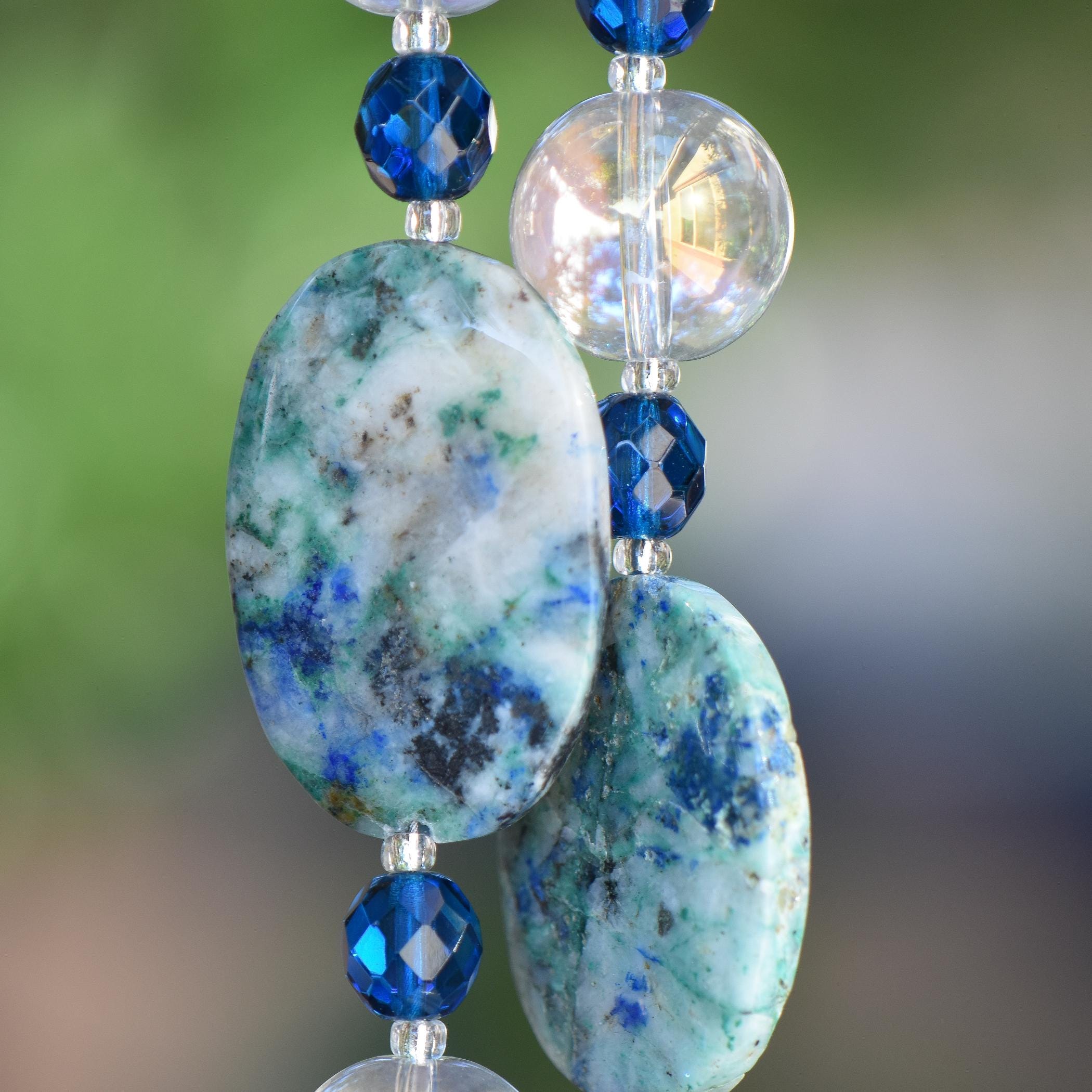 Close-up of a beaded wind chime featuring large, oval-shaped stones with a blue and green marbled pattern. The stones are interspersed with faceted, deep blue beads and clear, round beads. The background is a soft, blurred green.