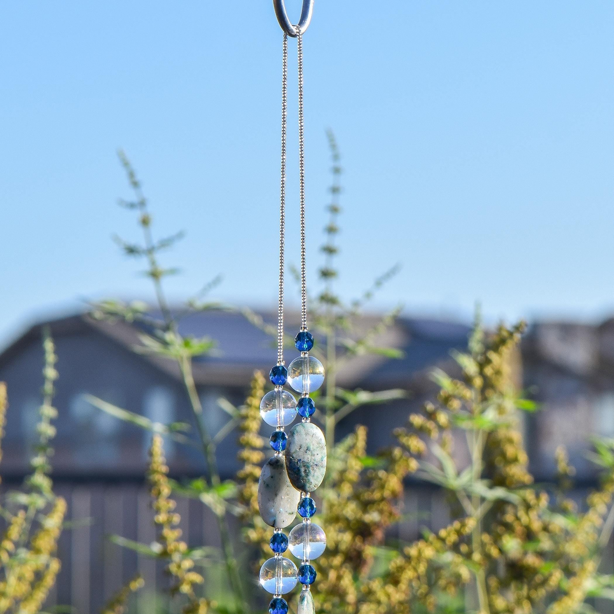 A decorative wind chime with a wire covered in silver-lined seed beads with blue and clear beads. The wind chime features a series of blue glass beads and oval-shaped stones in shades of blue and gray. The beads and stones are strung together, creating a visually appealing and elegant design.