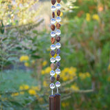 Bottom of Cinjo Handmade wind chime hanging in front of blurred foliage background. Two piece of transparent pale purple fused glass, rectangular-shaped, are anchors for two strings of beaded wire. Rhyolite stone beads are tear drop shaped and there is a larger, clear, reflective glass bead in between each stone bead. The stone beads are marked and in shades of brown, tan and gray.