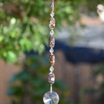 Five large oval Fire Opal stone beads strung with small glass crystal beads, anchored by large glass crystal prism shaped like the sun. Hanging vertically against blurred landscape background. Fire Opal beads have bits of opal stone surround by natural material that are mined with the gemstone and are neutral colors such as brown, tan and gray.