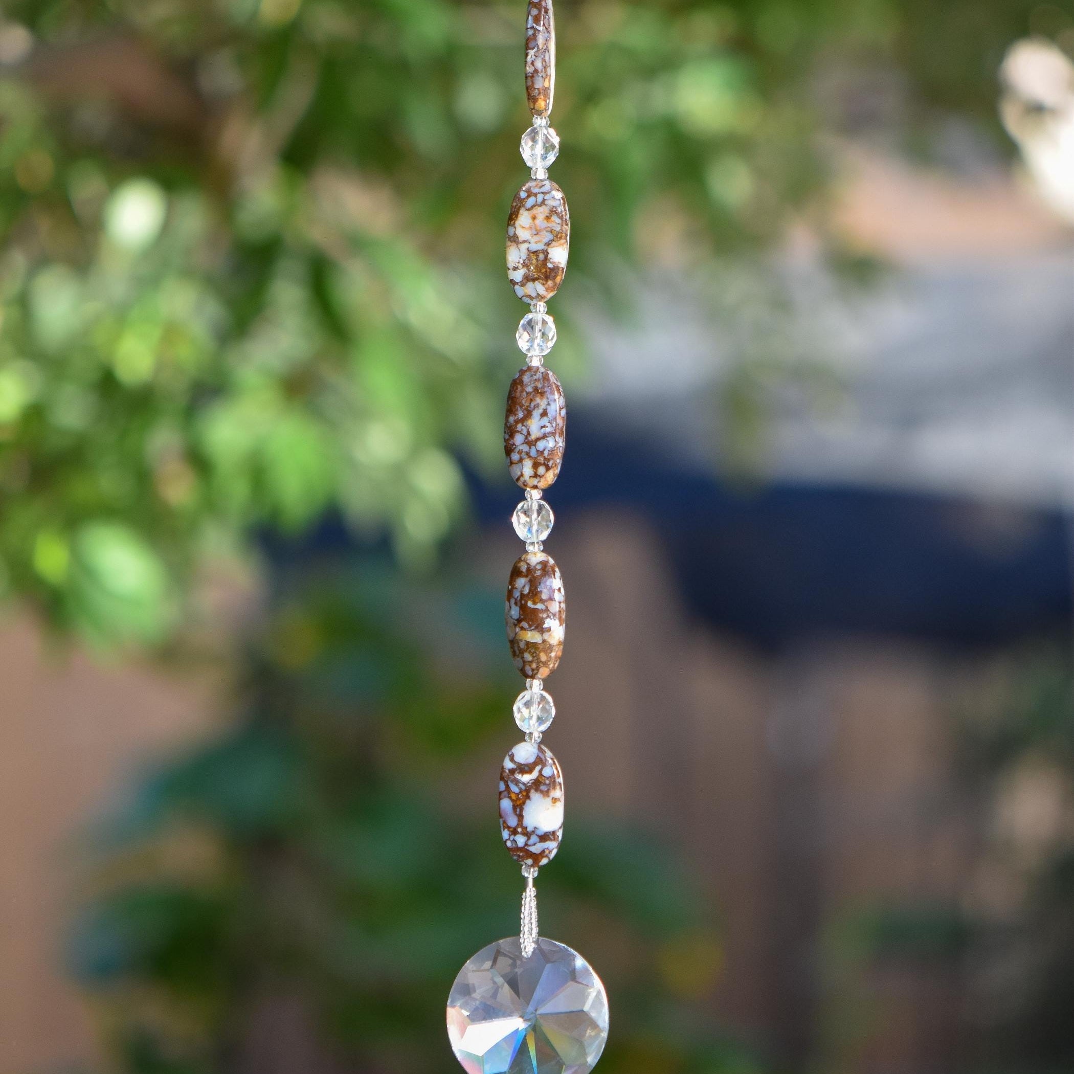 Five large oval Fire Opal stone beads strung with small glass crystal beads, anchored by large glass crystal prism shaped like the sun. Hanging vertically against blurred landscape background. Fire Opal beads have bits of opal stone surround by natural material that are mined with the gemstone and are neutral colors such as brown, tan and gray.