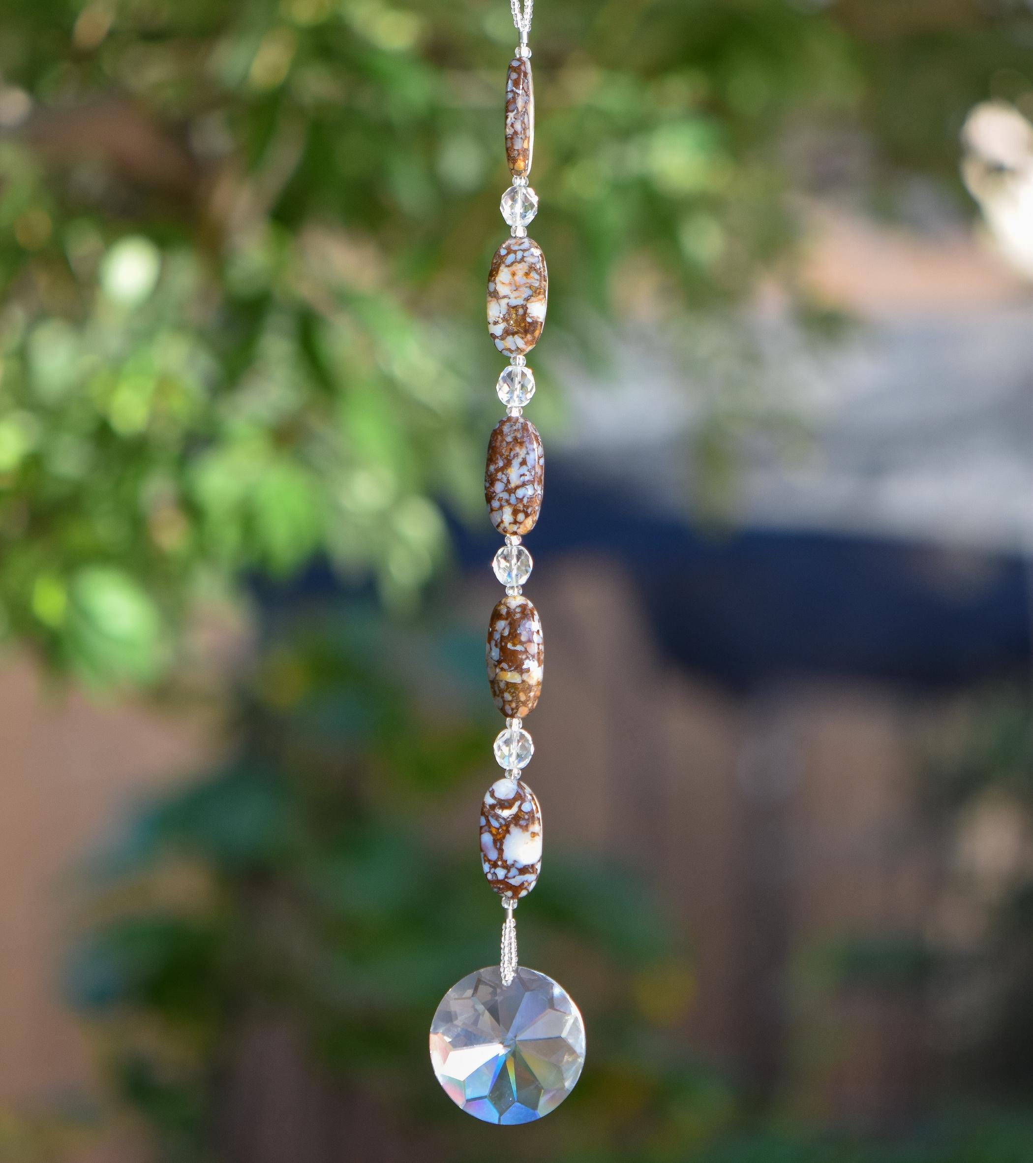 Five large oval Fire Opal stone beads strung with small glass crystal beads, anchored by large glass crystal prism shaped like the sun. Hanging vertically against blurred landscape background. Fire Opal beads have bits of opal stone surround by natural material that are mined with the gemstone and are neutral colors such as brown, tan and gray.
